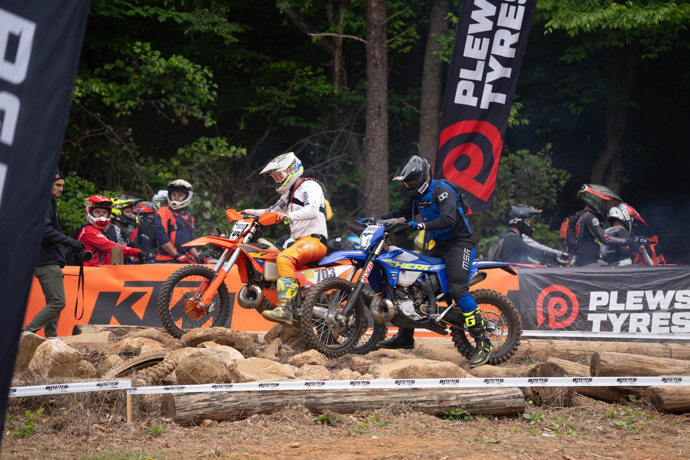 Two hard enduro racers on an orange KTM and a blue Sherco at a log and rock start line as other riders wait behind