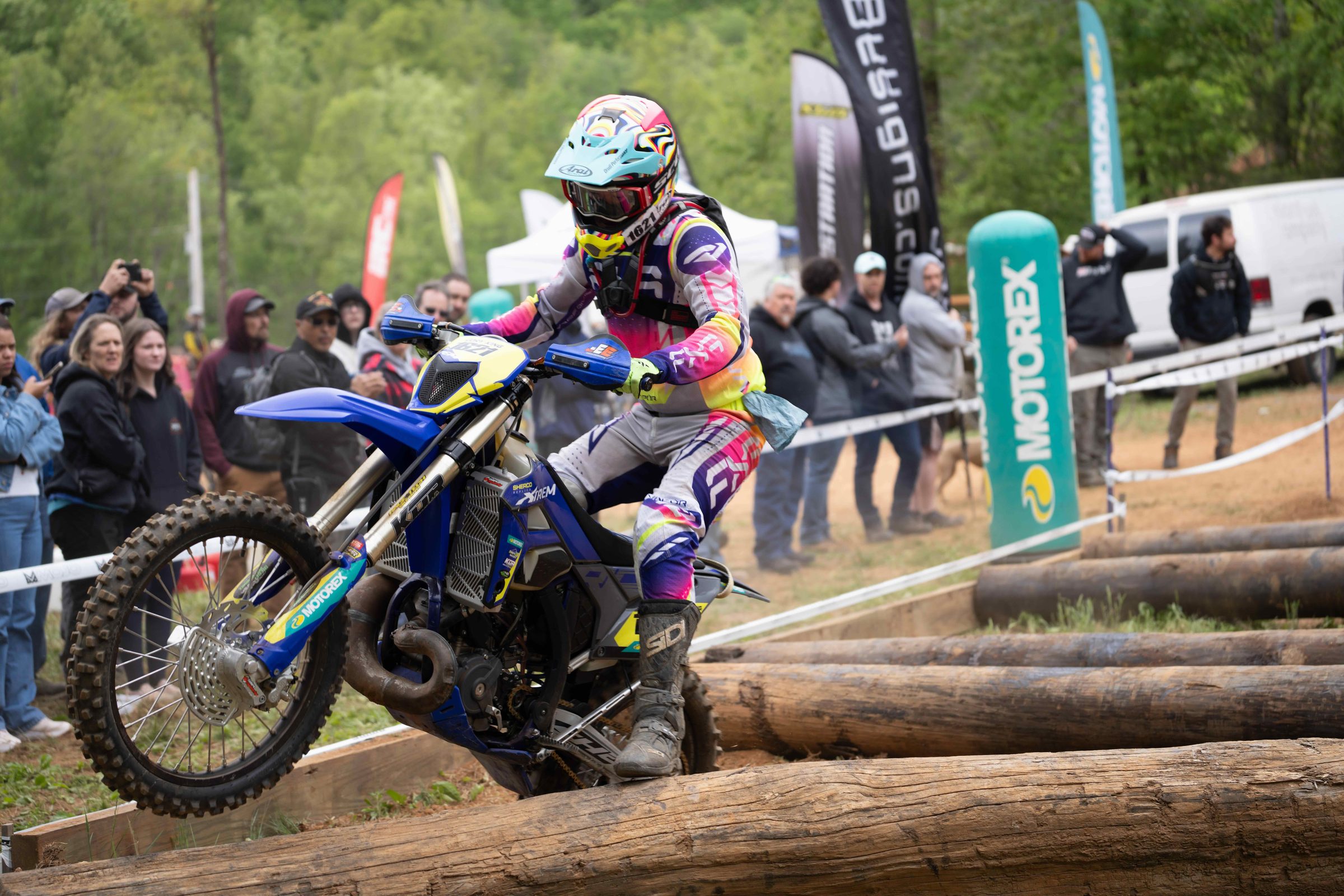 Hard enduro racer in bright gear lifting the front wheel over a log crossing as the crowd watches